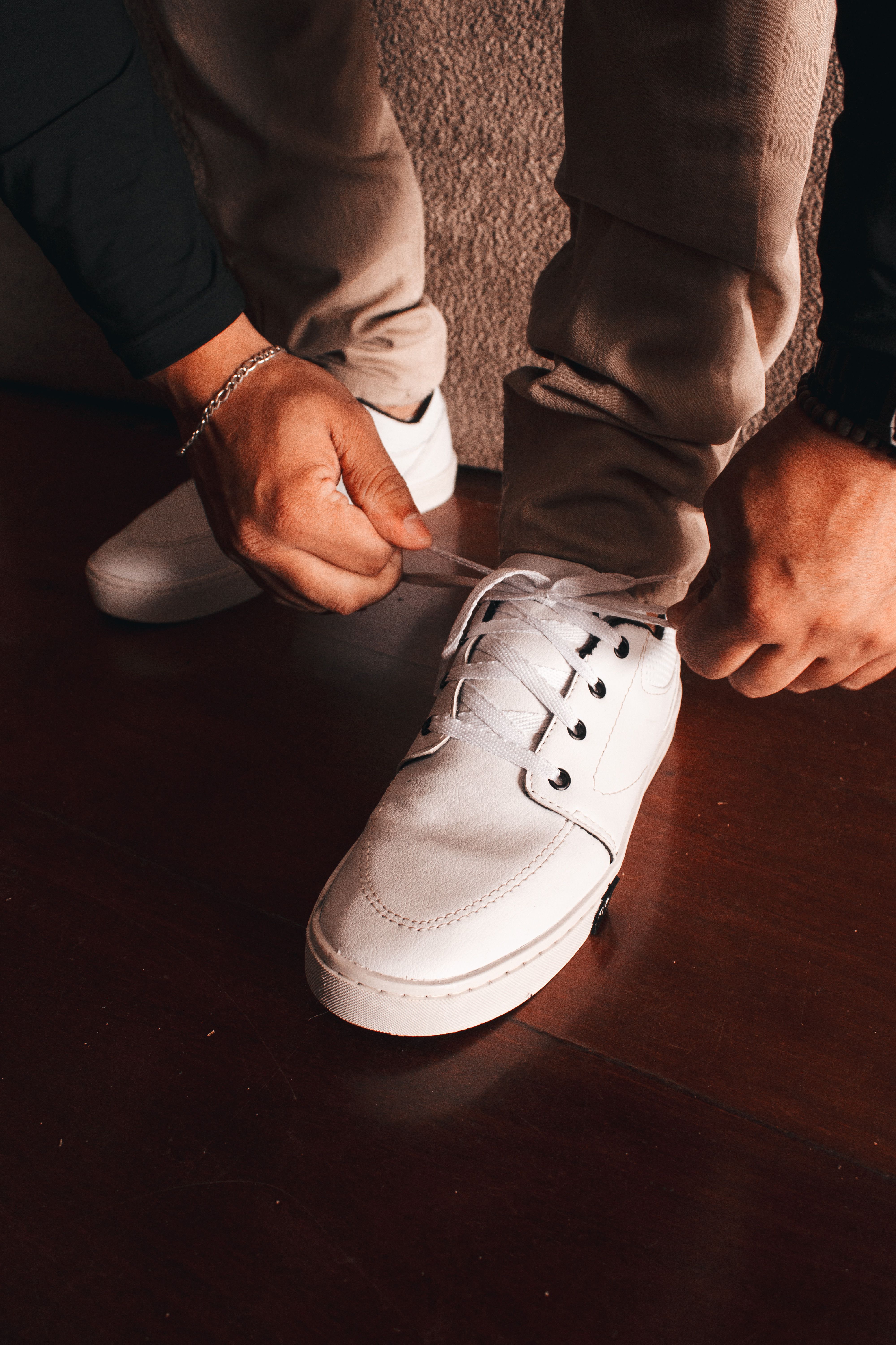 Close-up of a man tying white shoes on wooden floor indoors.