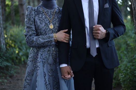 Couple in formal attire with intricate details on gown holding hands in a serene outdoor setting.