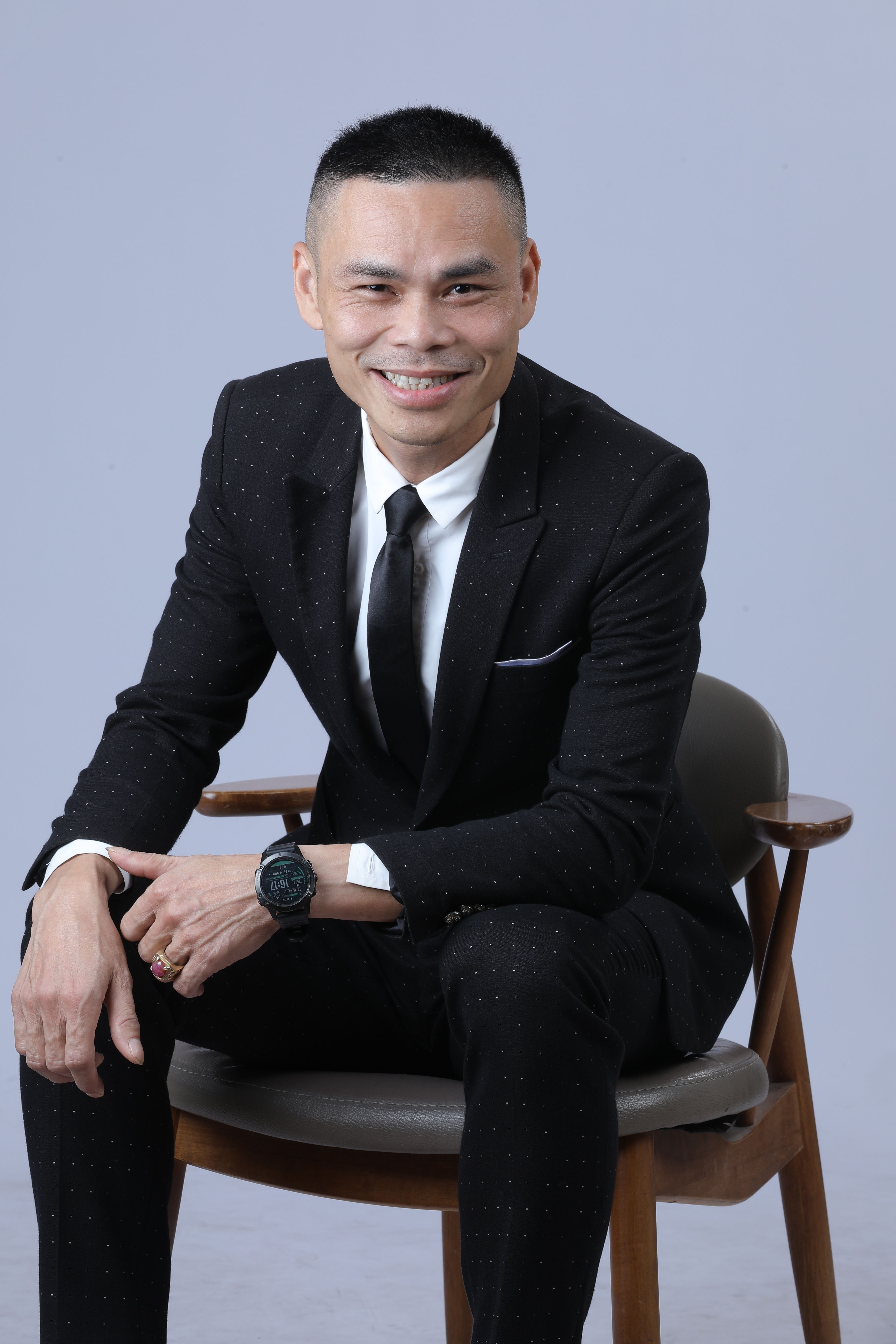 Asian man in a stylish black suit sitting on a chair, smiling warmly.