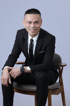 Asian man in a stylish black suit sitting on a chair, smiling warmly.
