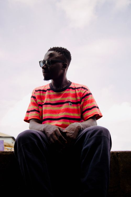 Free A young man wearing sunglasses and a striped shirt sits outdoors under a cloudy sky. Stock Photo