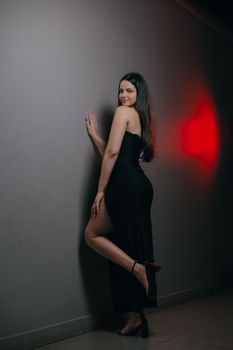 A woman in a black dress poses gracefully against a gray wall with a red light accent.