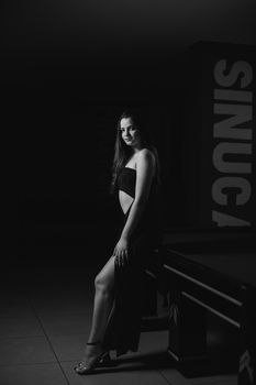 Stylish woman in black dress poses by a pool table, showcasing elegance in monochrome.