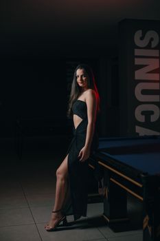 A young woman in a black dress poses elegantly beside a pool table indoors.