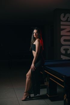 Beautiful woman posing in black evening dress next to a pool table indoors.