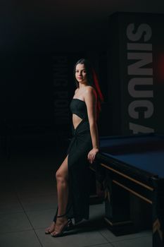 Young woman in a glamorous black dress posing elegantly by a pool table indoors.