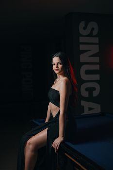 A stylish young woman in a black dress poses elegantly on a pool table indoors.
