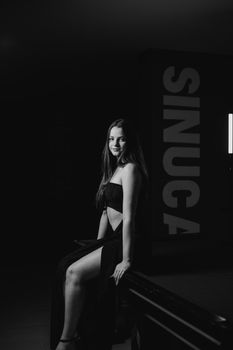A fashionable young woman poses elegantly in a black dress on a pool table in a dark room.