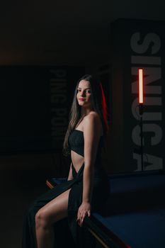 A young woman in a black dress elegantly poses on a pool table with moody lighting.