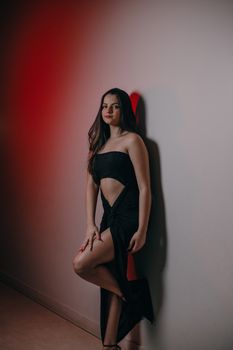 A young woman in an elegant black dress poses against a wall in a studio setting.