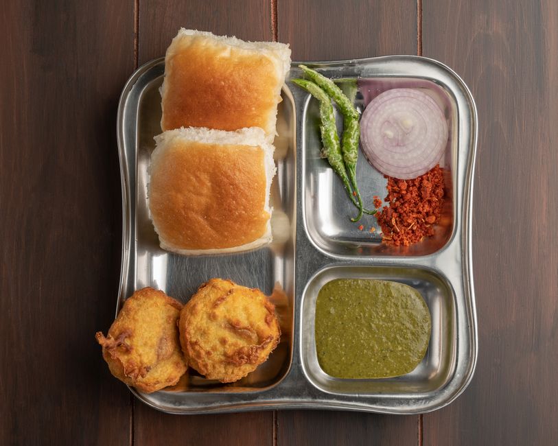 A vada pav sandwich cut open showing the spiced potato vada inside a soft pav bun, smeared with red and green chutney, served with a fried green chili on the side.
