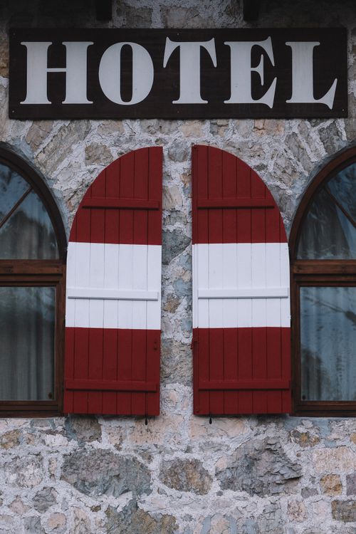 Free Close-up of a stone hotel facade with red and white wooden shutters, creating a cozy and inviting atmosphere. Stock Photo