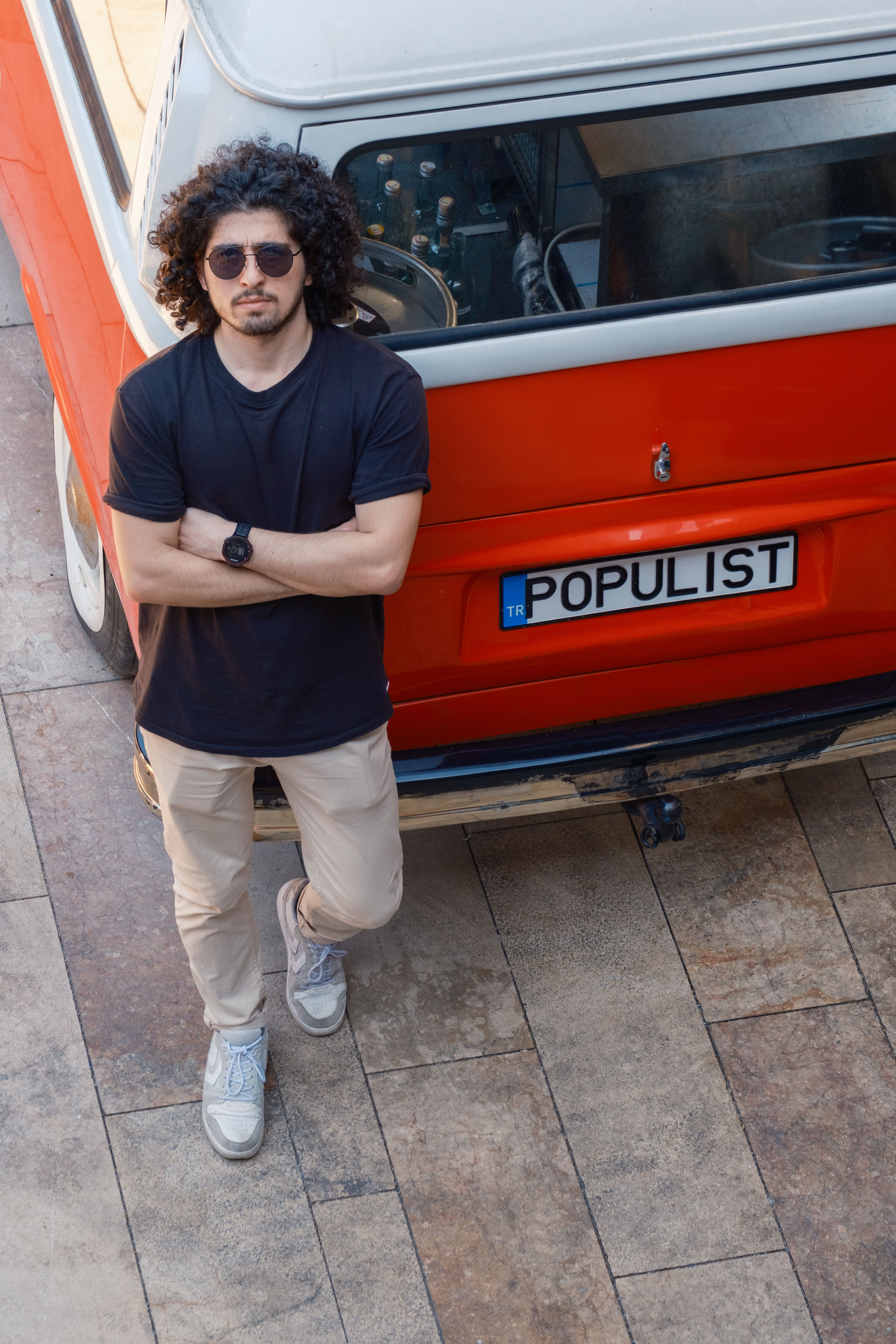 Young man with sunglasses and curly hair leaning on a red vintage Volkswagen minibus wearing casual attire.