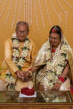 Senior couple in traditional attire cutting a celebratory cake adorned with flower garlands.