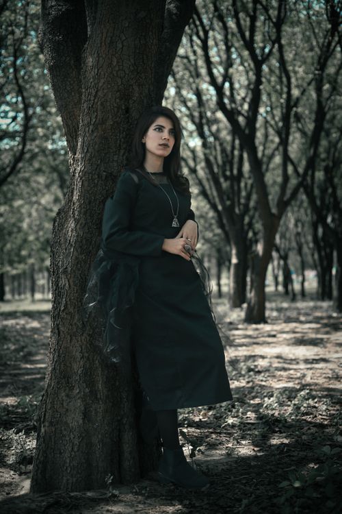Free Portrait of a fashionable woman in dark dress leaning against a tree in a serene forest in Islamabad. Stock Photo