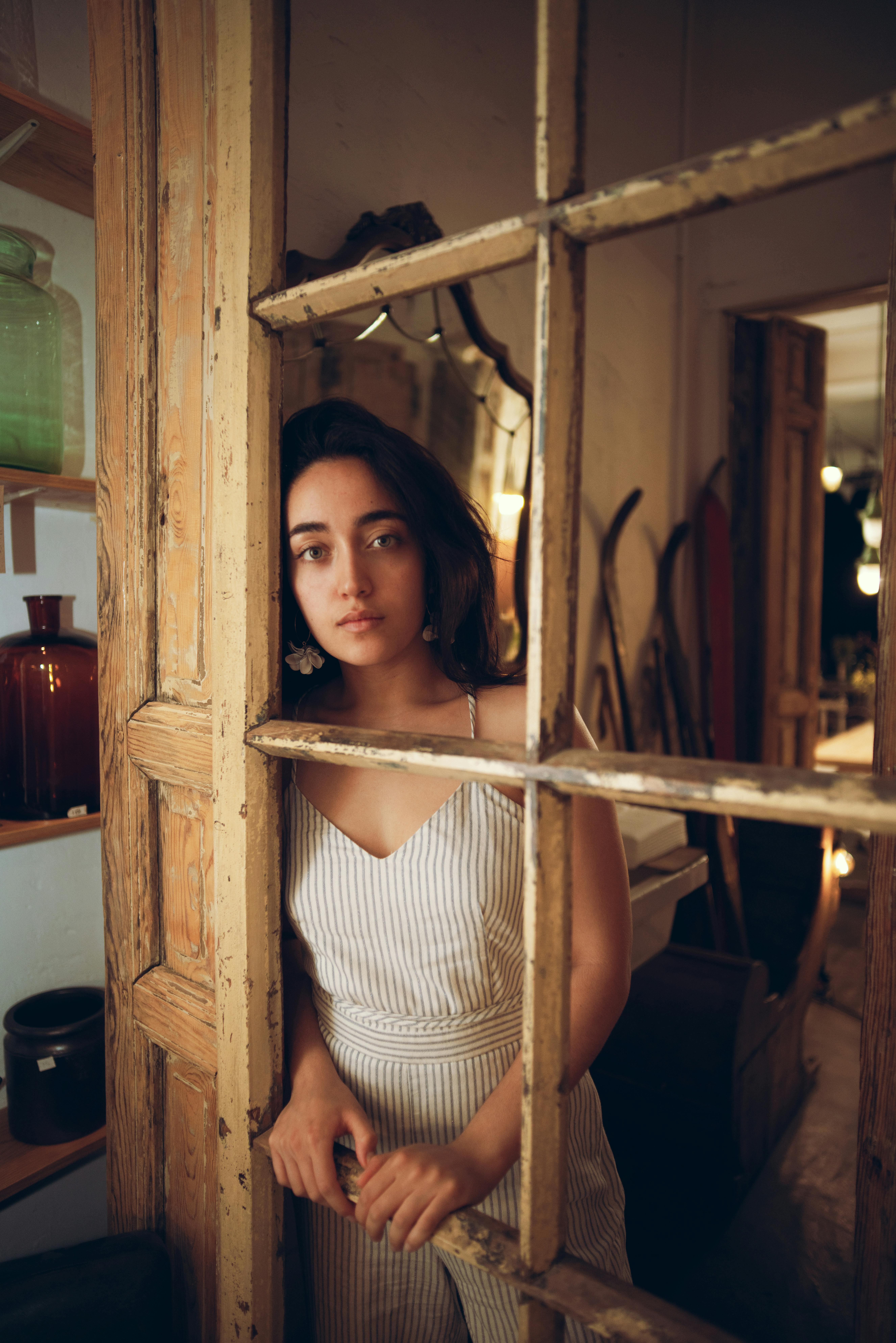 A young woman with brunette hair standing indoors by a vintage wooden doorway, looking thoughtful.