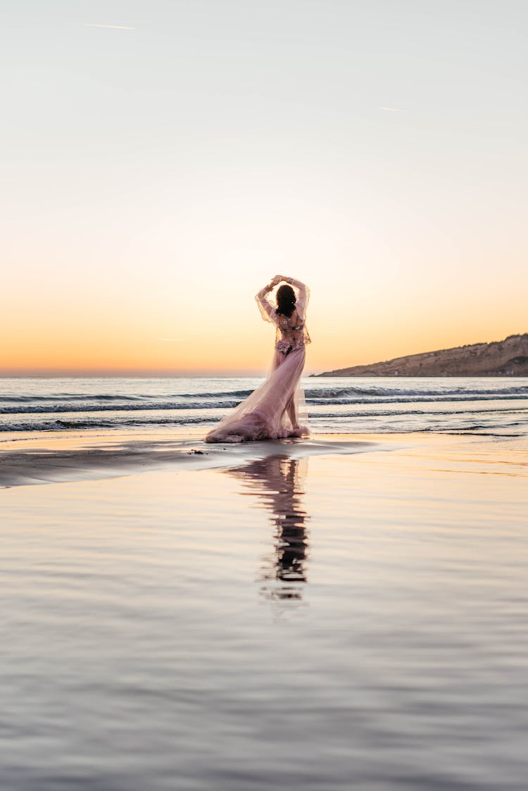 Woman In A Long Pink Dress Wading In The Sea On The Beach At Sunset