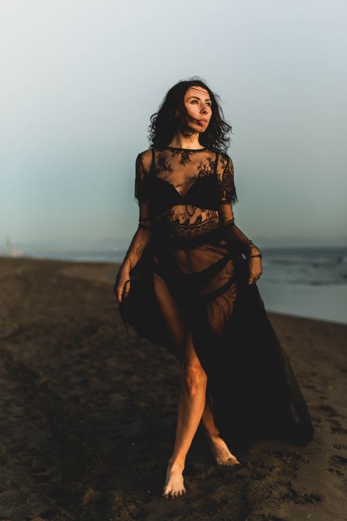 Free A woman in a sheer black dress walking along a serene beach at sunset. Stock Photo