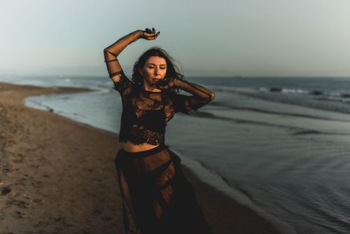 Free Artistic portrait of a woman in a black veil dress posing on a tranquil beach. Stock Photo
