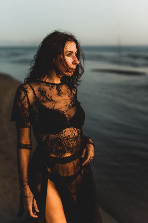 Free A stylish woman in black lace near the ocean shore at sunset. Captivating evening scene. Stock Photo