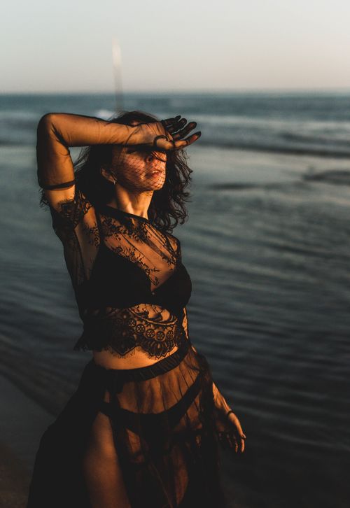 Free A woman in a black lace dress poses elegantly by the beach at sunset. Stock Photo