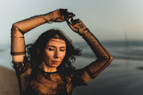 Brunette Woman in Black Lace Dress, Mesh Gloves and Veil Posing at a Sea Beach