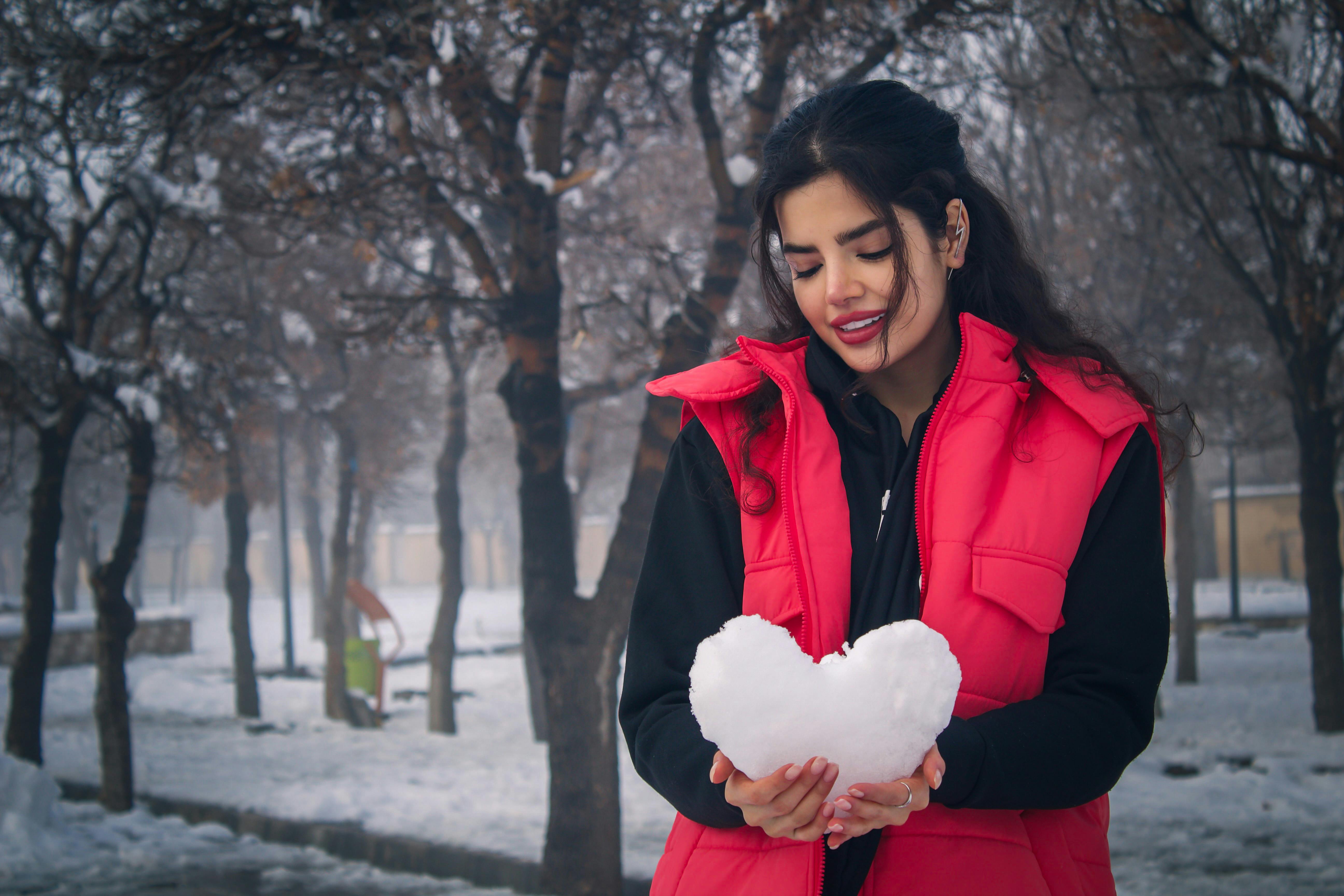 A woman in a red jacket holding a heart-shaped snow outdoors in a snowy park setting.