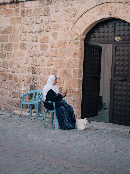 Free An elderly woman in a headscarf sits outside a stone building with a large entrance door. Stock Photo
