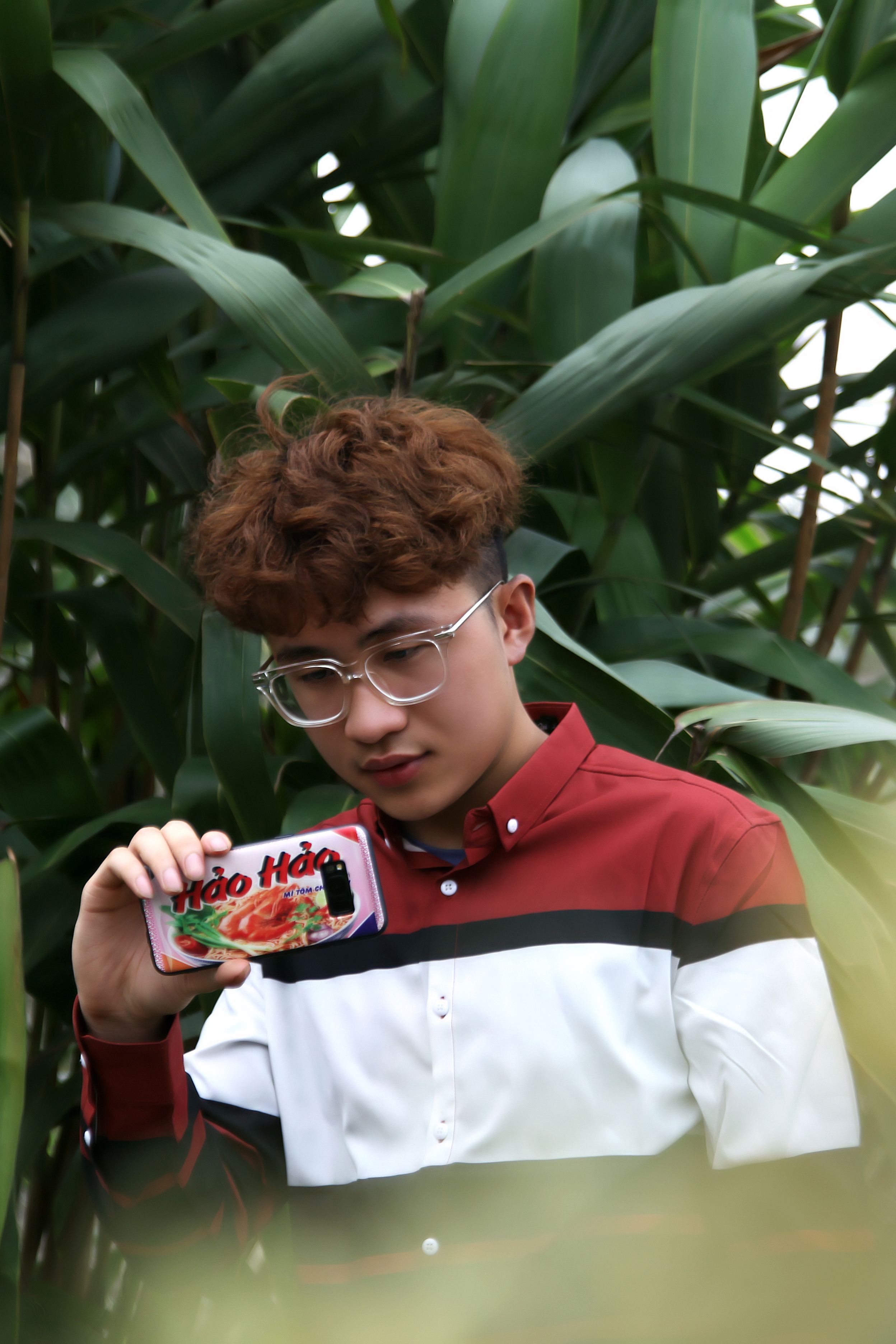 Fashionable young man with glasses taking a photo using a smartphone in a lush park.