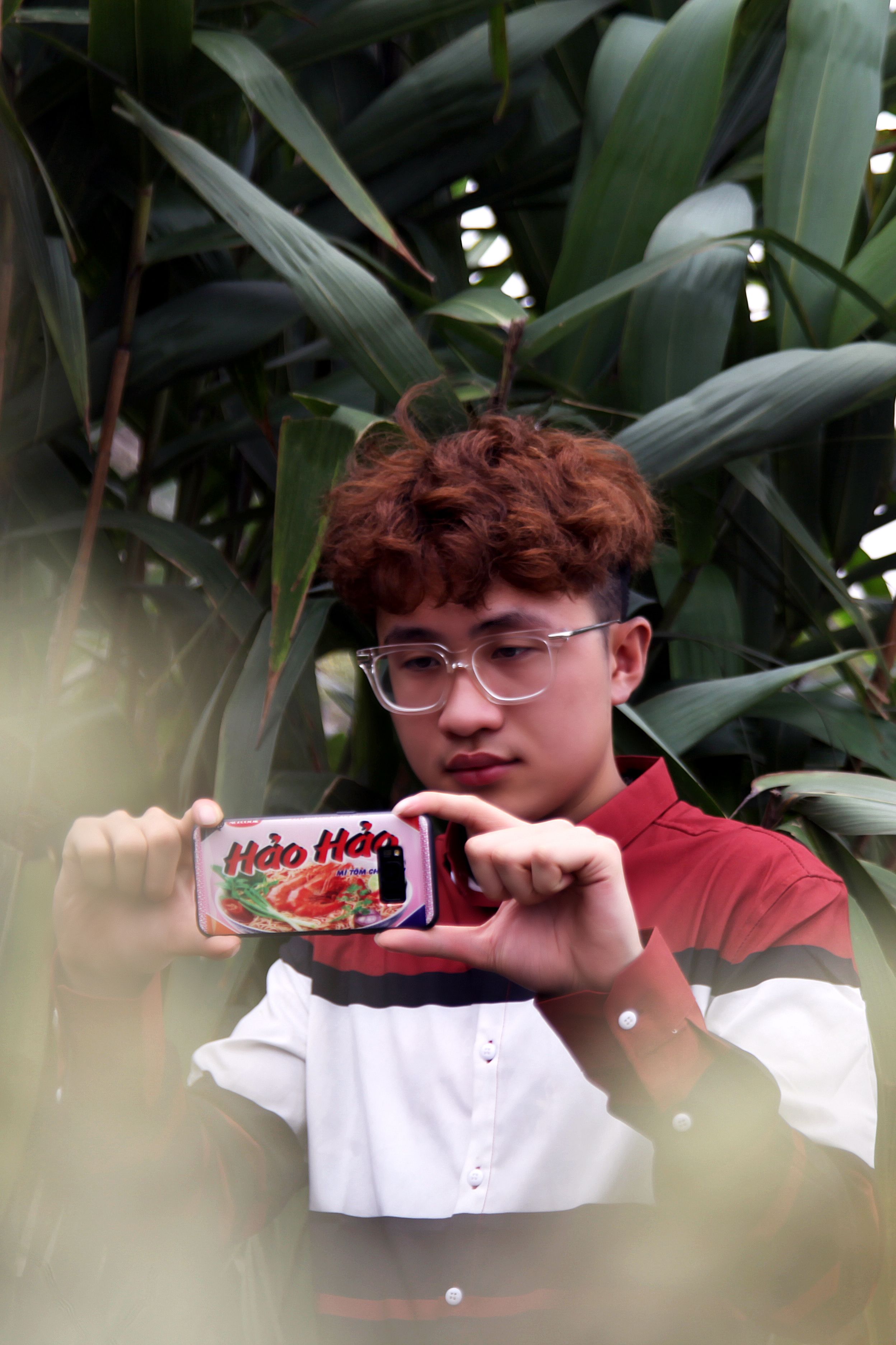 A teenager with dyed hair takes photos using a smartphone amidst lush greenery.