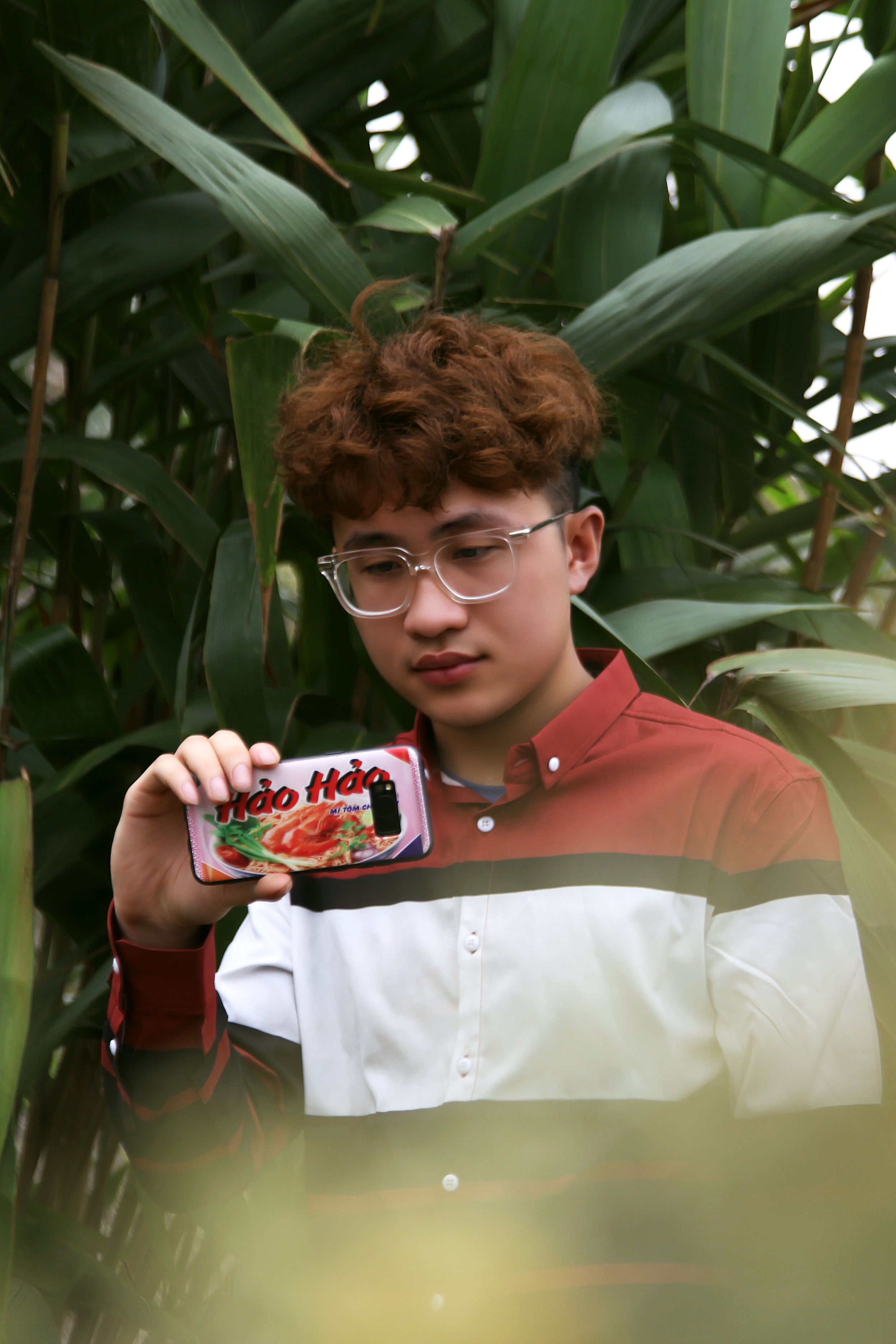 Young Asian male holding snack pack in lush tropical garden, wearing glasses and a striped shirt.