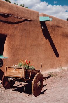 Sunlit rustic wooden cart with flowers beside an orange adobe wall under a blue sky.