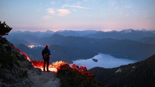 A Man with a Backpack and Flashlight Standing on a Trail overlooking a Lake and Mountains at Sunset