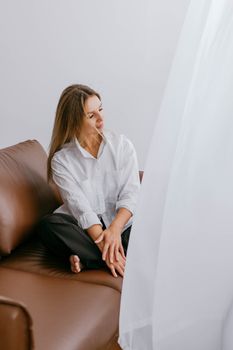 A woman in a white shirt sitting on a brown sofa, looking towards a curtain, creates a serene indoor setting.