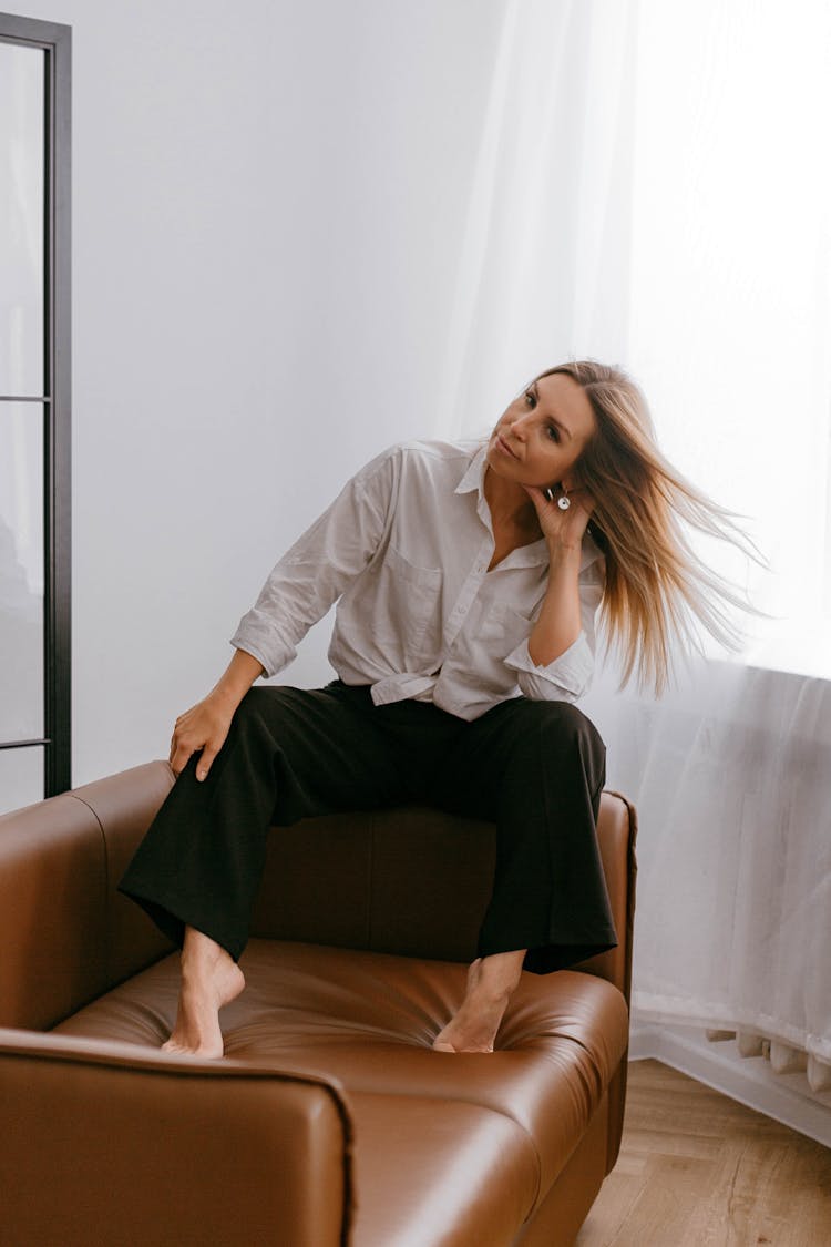 Blonde Woman In White Blouse And Black Wide Trousers Sitting On A Leather Sofa