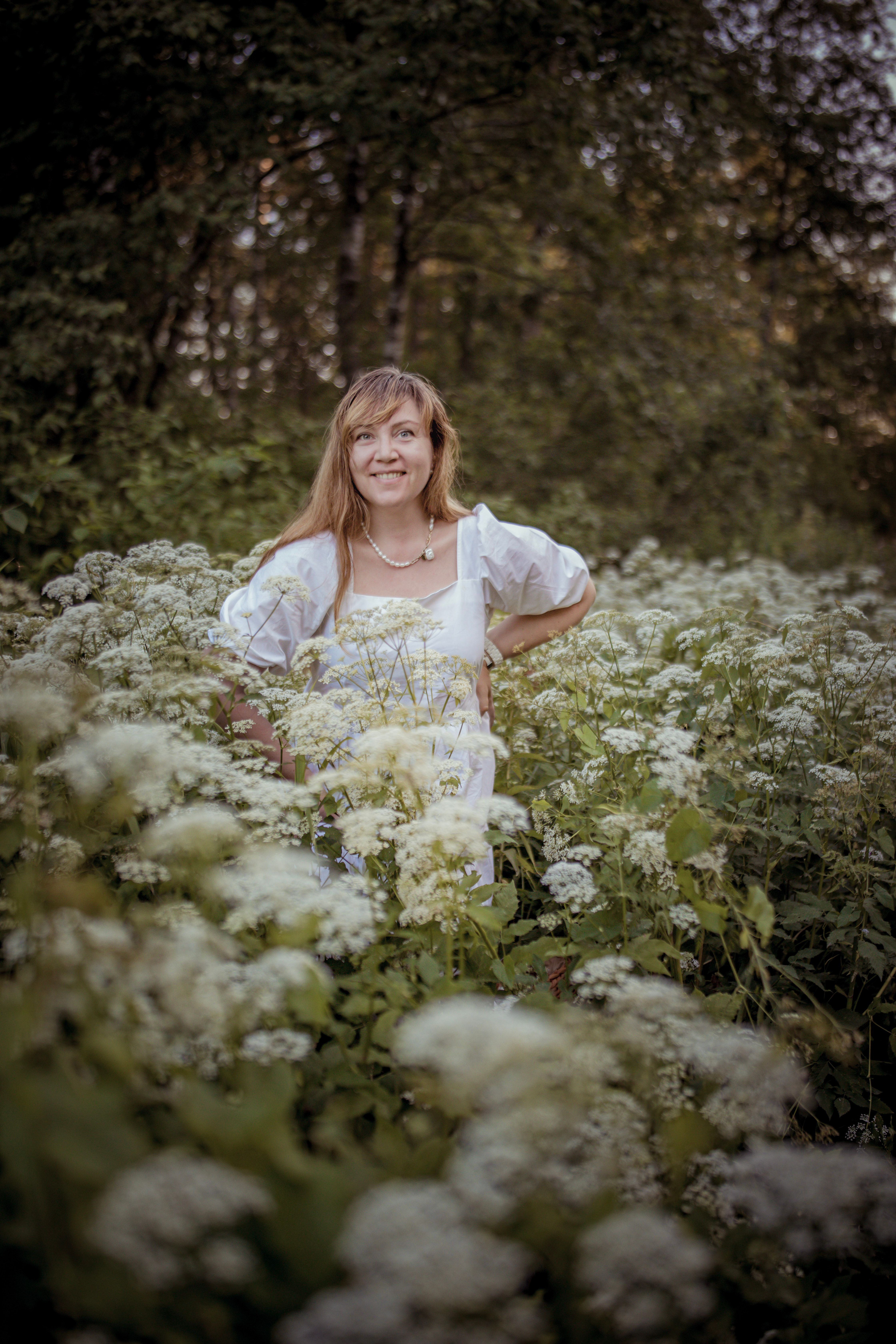 A woman smiling in a lush meadow filled with white wildflowers on a sunny day.