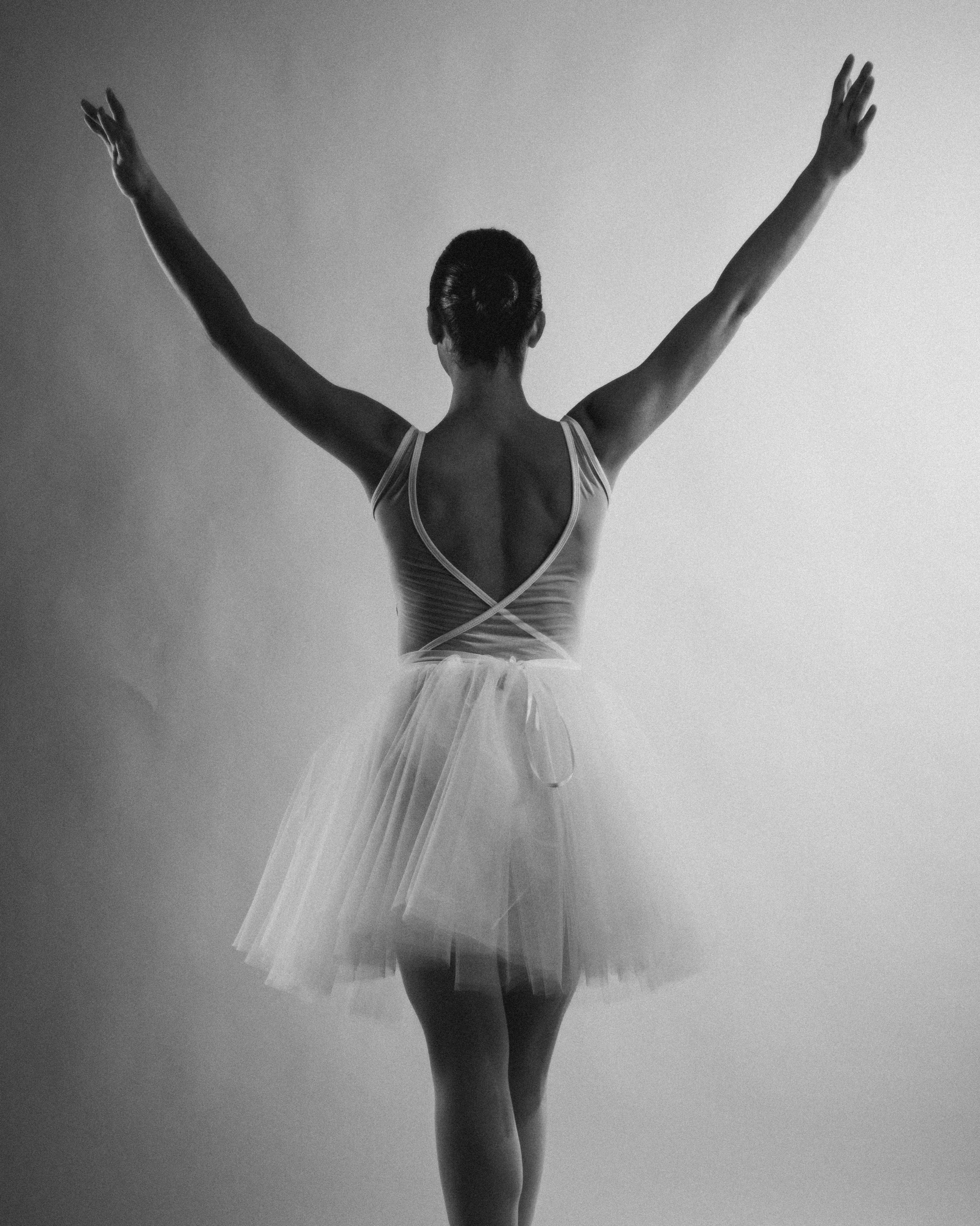 Free A graceful ballerina in a tutu striking an elegant pose with arms raised, back view in black and white. Stock Photo