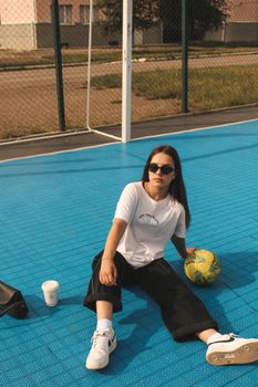 Stylish woman in sunglasses sitting on a sunlit basketball court with a ball.