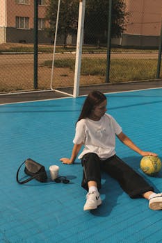 A young woman sits on a blue sports court with a ball, bag, and drink, enjoying a sunny day.