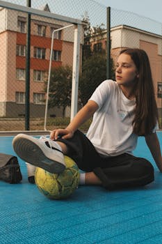 Teenage girl in casual sportswear sitting on urban soccer field with a ball.