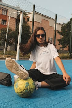 Teen girl sitting with soccer ball on urban field, casual style.