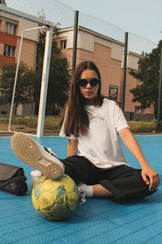Stylish young woman sitting on a basketball court with a football, wearing sunglasses.
