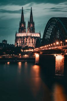 Stunning night view of Cologne Cathedral and Hohenzollern Bridge with city lights reflecting on the Rhine River.