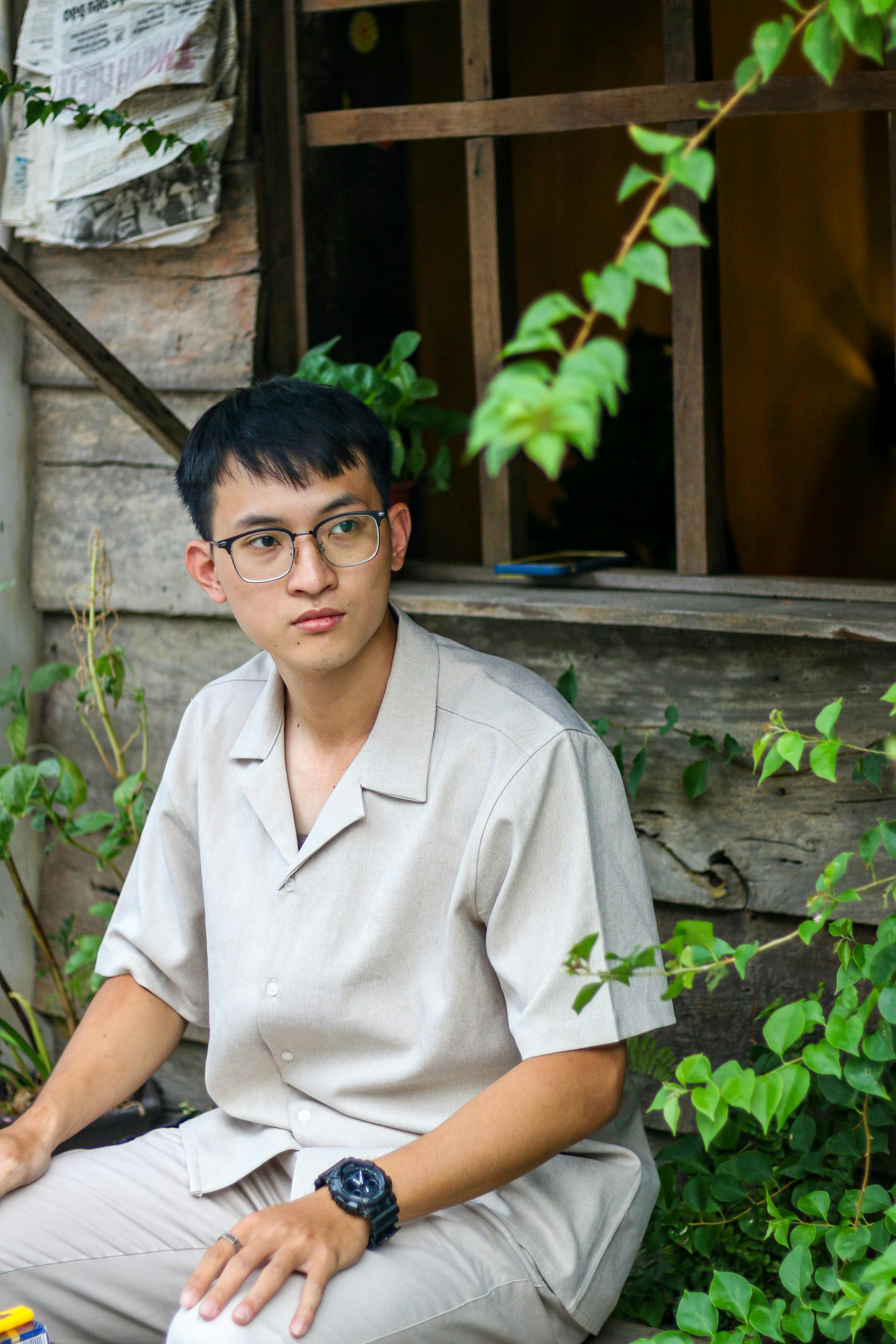 A young Asian man sits by a rustic wooden house surrounded by greenery, wearing eyeglasses and a casual shirt.
