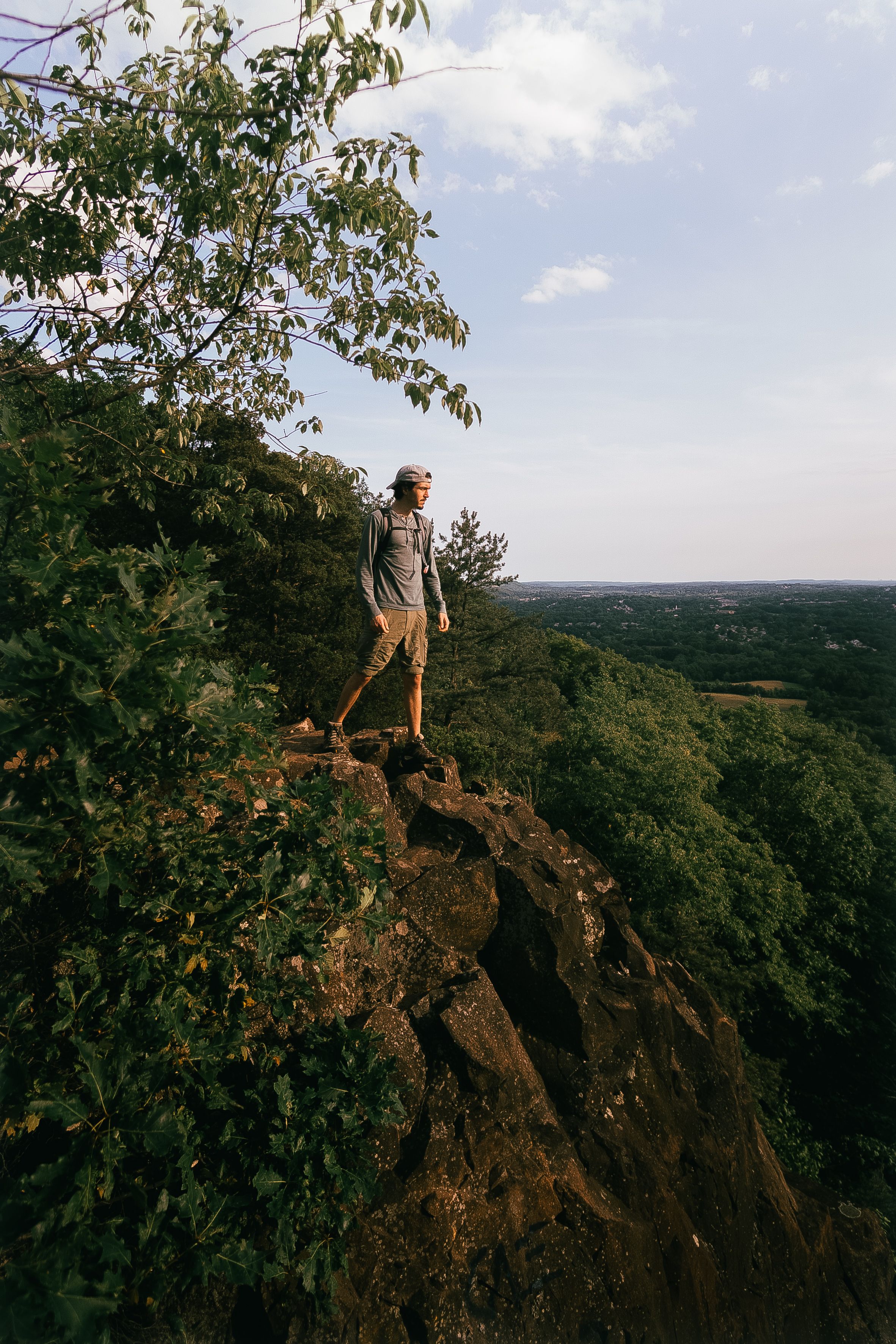Man stands on cliff edge overlooking forest landscape, embracing nature and adventure.