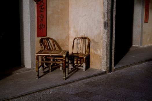 Bamboo chairs and table against rustic wall with sunlight and traditional decor in quiet outdoor setting.