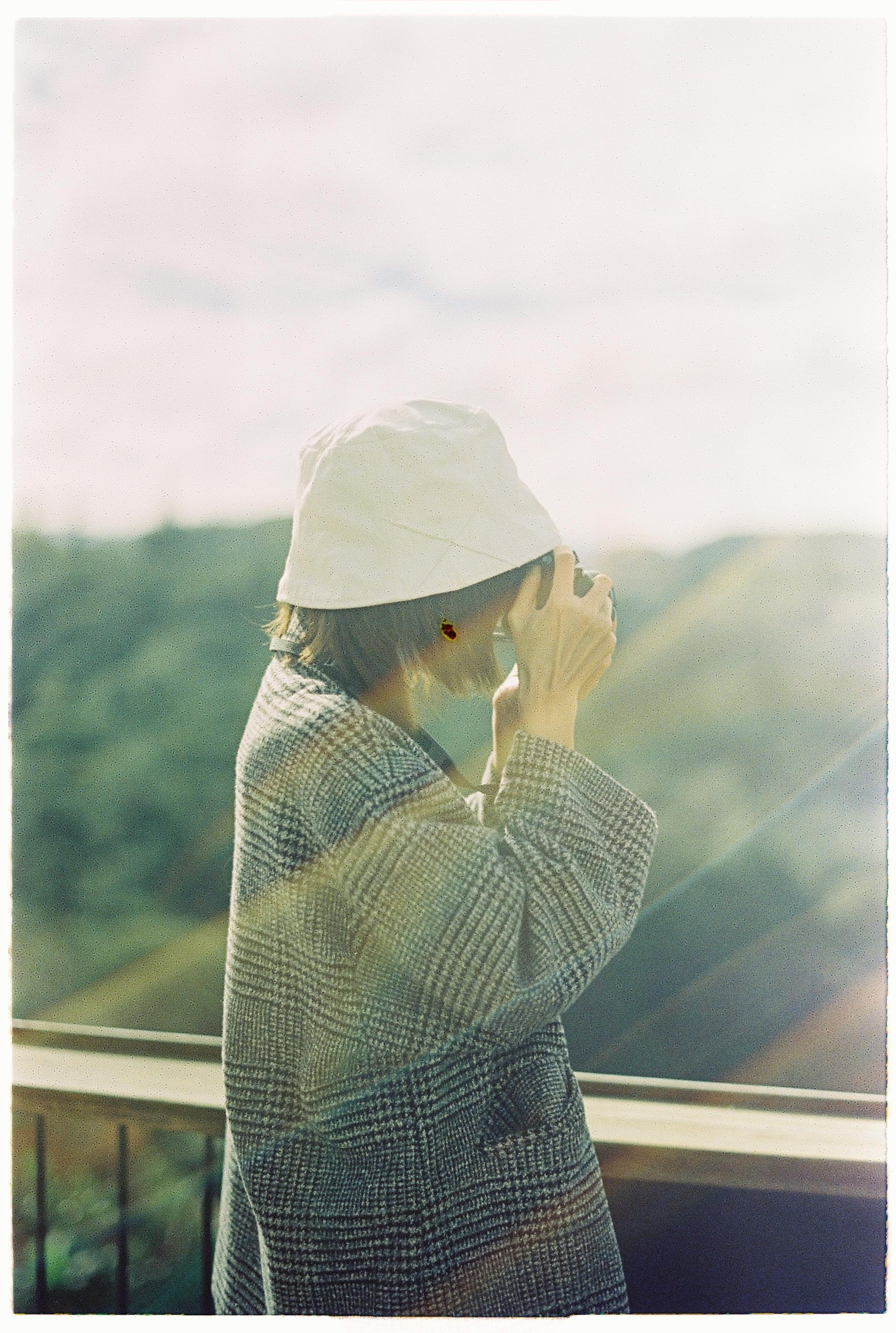 A woman wearing a hat and coat takes a picture outdoors with striking lens flare.