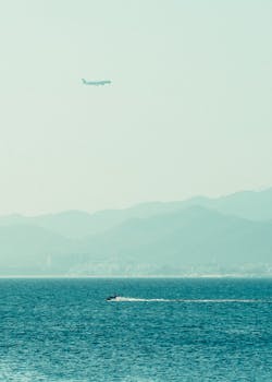 Airplane soaring above tranquil sea with a boat, captured against a misty mountain backdrop.