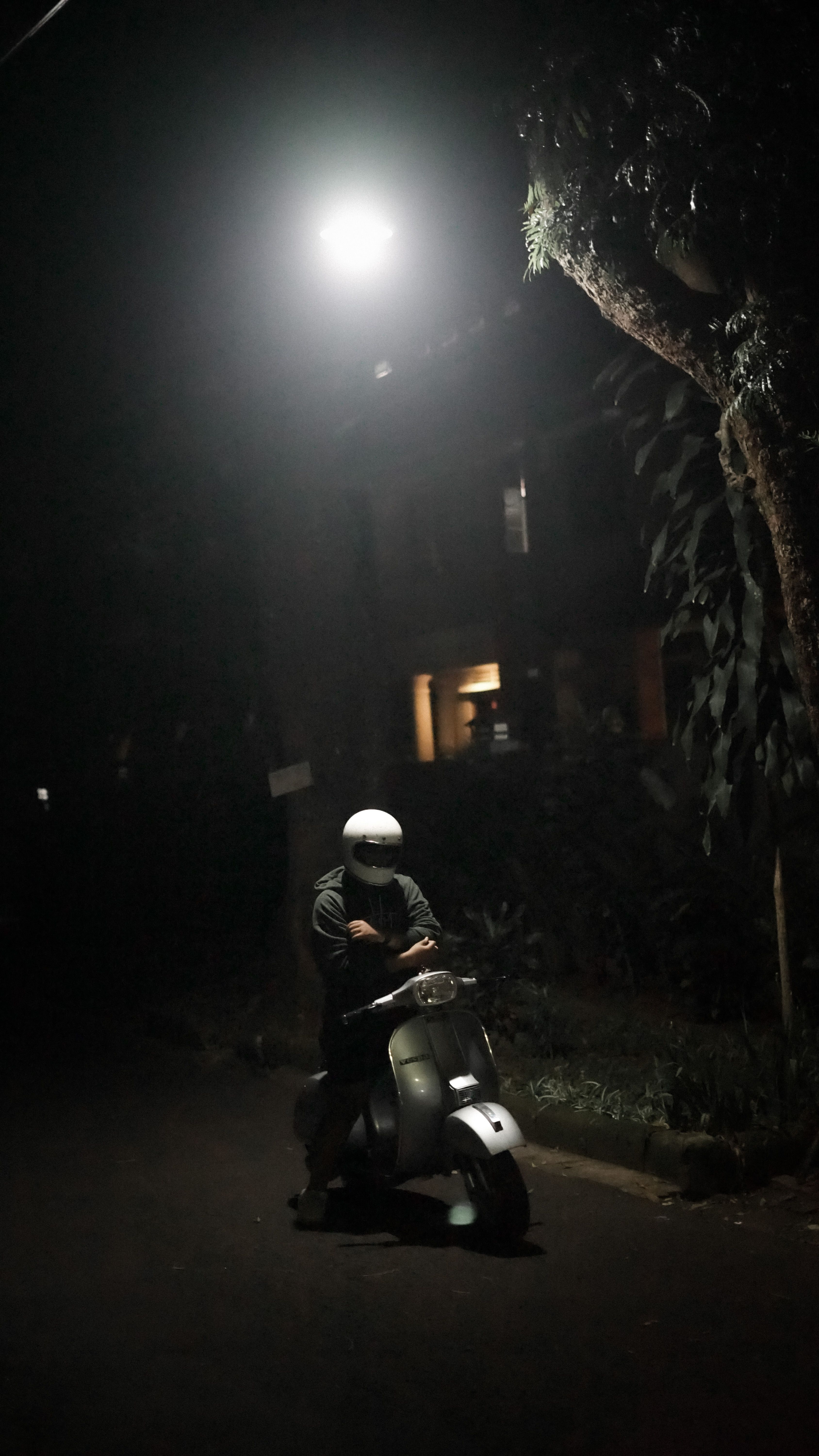 A helmeted person stands next to a motor scooter on a dimly lit urban street at night.