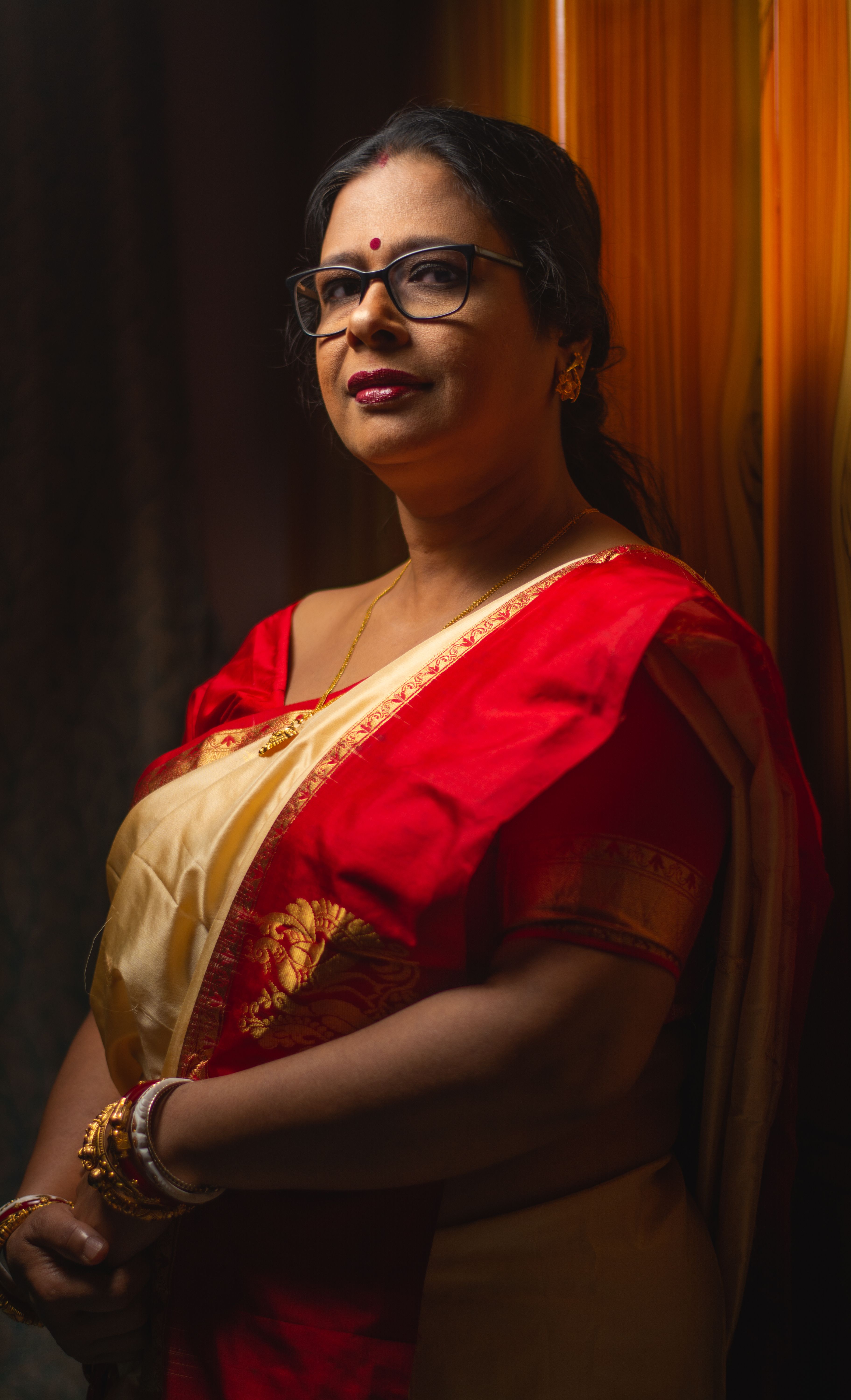 A woman in eyeglasses wearing a traditional red and gold sari with a warm smile indoors.
